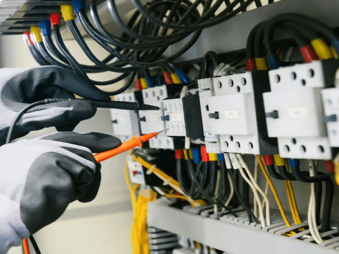 Teknisyenin eldivenli elleri multimetre ile elektrik panosunu test ediyor.
